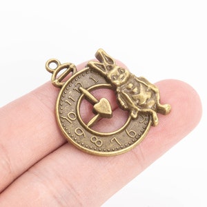 May include: A bronze-colored charm featuring a rabbit holding a pocket watch with Roman numerals. The rabbit is sitting on the watch face, which has a heart-shaped design in the center.