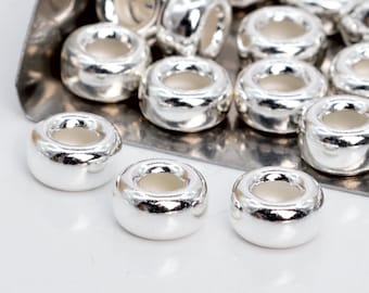 Può includere: Una collezione di perline lucide color argento a forma di ciambella. Le perline hanno una superficie liscia e riflettente e un foro al centro. Sono esposte su un vassoio d'argento e su una superficie bianca.