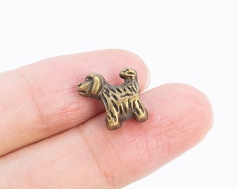 May include: A small, gold-colored metal charm in the shape of a dog. The charm is detailed with a textured surface and is being held by a finger.