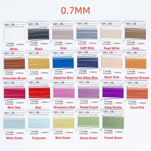 May include: A collection of silk thread spools in various colors, including white, black, gray, and shades of blue, pink, and green. Each spool is labeled with its color and includes a needle and 2 meters of thread. The text "0.7MM" is at the top.