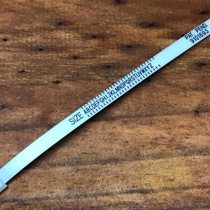 May include: A white plastic ring sizer with black markings. The sizer has a size chart with letters A through Z and the text "SIZE" printed on it. The sizer also has the text "PAT. PEND. 9101693" printed on it.