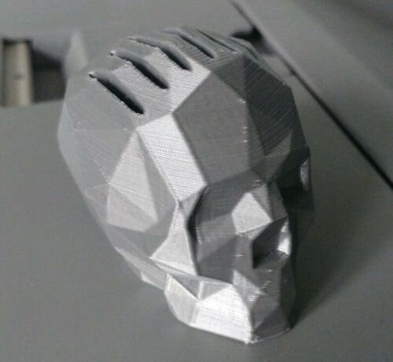 May include: A 3D printed skull sculpture made of gray plastic. The skull is designed in a geometric style with a faceted surface.