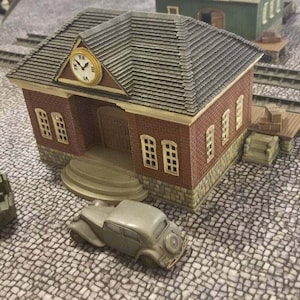 May include: A miniature model of a brick building with a clock tower and a grey tiled roof. The building has a white car parked in front of it. The building is on a grey cobblestone surface.