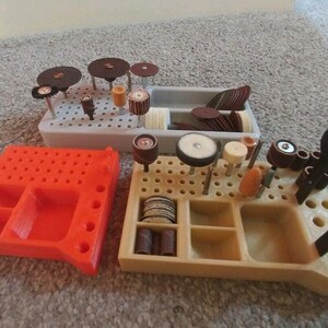 May include: Three 3D printed organisers for storing small tools and accessories. The organisers are made of orange, beige and grey plastic. The beige organiser has a variety of small tools and accessories stored in it, including sanding discs, drill bits and wrenches.