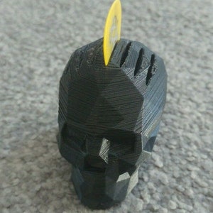 May include: A black 3D printed skull shaped guitar pick holder with a yellow guitar pick inserted in the top.