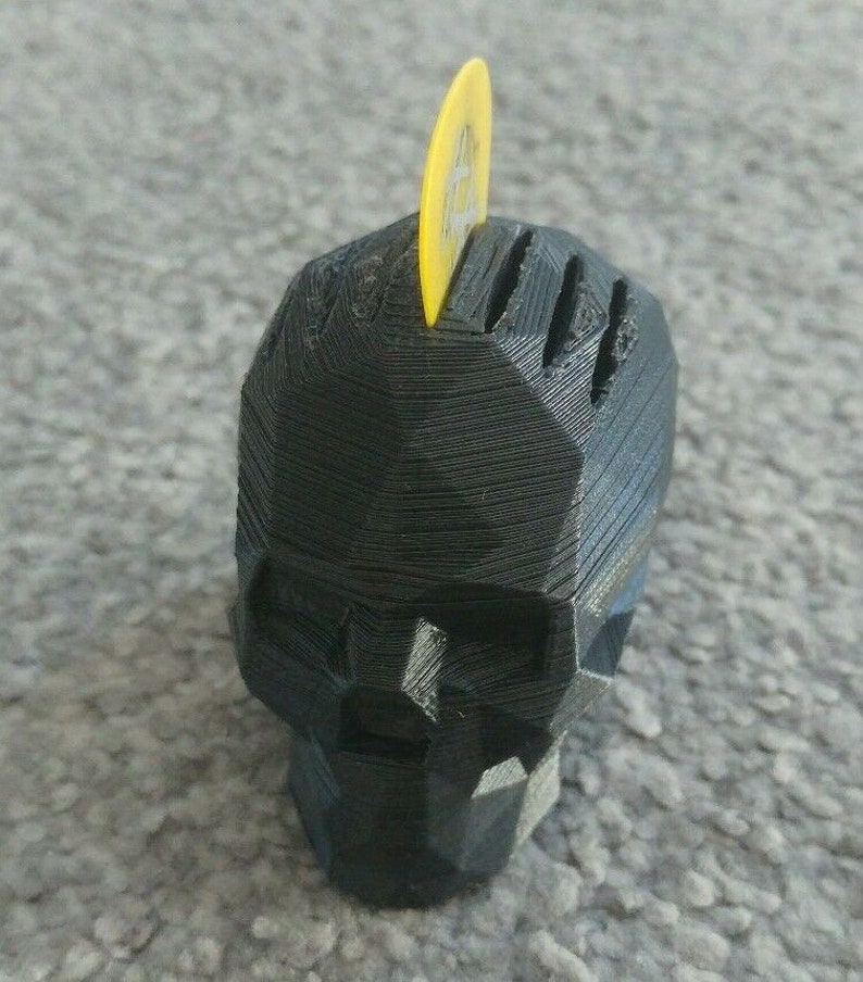 May include: A black 3D printed skull shaped guitar pick holder with a yellow guitar pick inserted in the top.