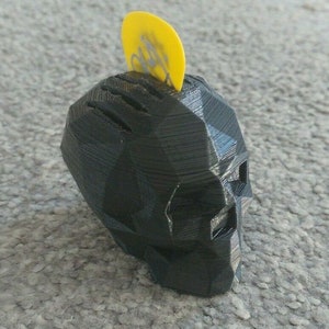 May include: A black 3D printed skull with a yellow guitar pick on top. The skull has a geometric design and is a unique and decorative item.