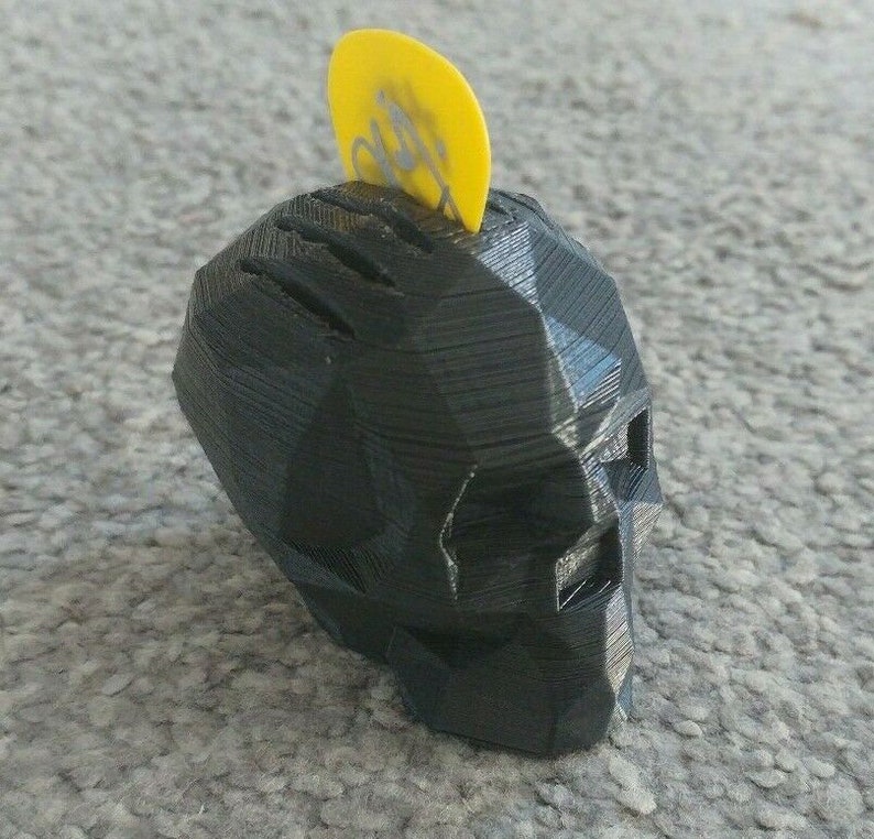 May include: A black 3D printed skull with a yellow guitar pick on top. The skull has a geometric design and is a unique and decorative item.