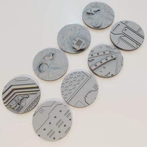 May include: Eight grey, circular miniature bases with diverse textured designs. Some bases feature raised mechanical details, while others have terrain-like textures. The bases are arranged on a white surface.