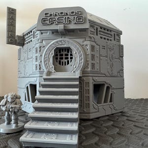 May include: A grey, 3D-printed model of a futuristic casino building with the text "CHRONOS CASINO" on the front. The building has a detailed design with stairs leading to the entrance and a small figure of a soldier.