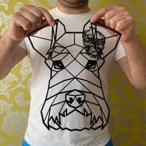May include: Black geometric dog head art piece. The artwork features a stylized dog face with a skull design on the right ear. The dog's face is made up of black lines and shapes. The art piece is held up by a person.
