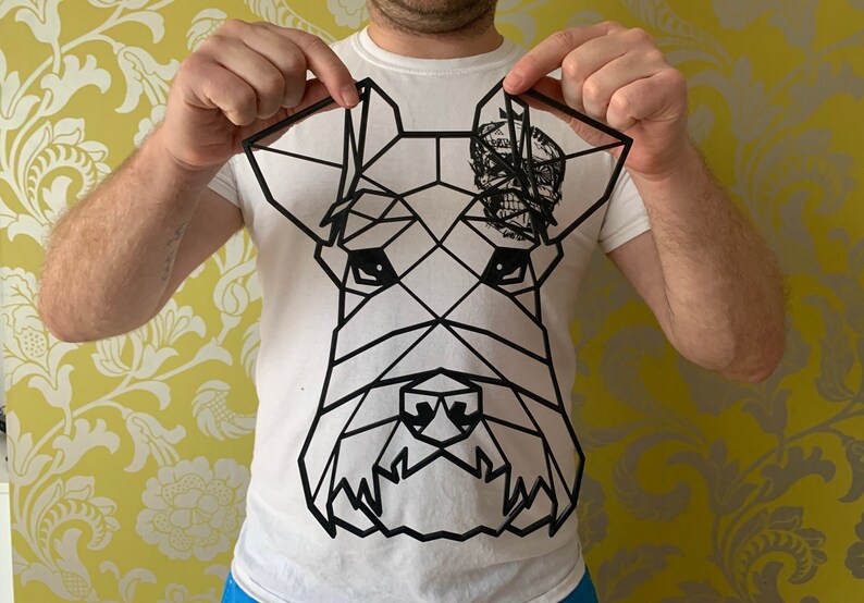 May include: Black geometric dog head art piece. The artwork features a stylized dog face with a skull design on the right ear. The dog's face is made up of black lines and shapes. The art piece is held up by a person.