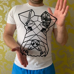 May include: A white t-shirt featuring a geometric black line drawing of a dog's face. The dog's ear has a patterned design. The person is holding the dog drawing in front of them.