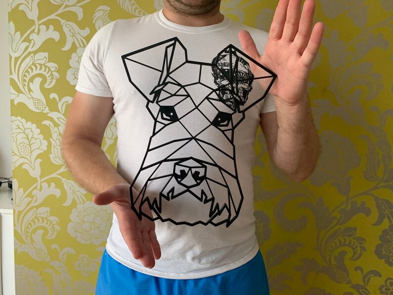 May include: A white t-shirt featuring a geometric black line drawing of a dog's face. The dog's ear has a patterned design. The person is holding the dog drawing in front of them.