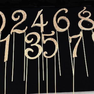 May include: Ten gold-colored cake toppers, each featuring a different number from 0 to 9, adorned with sparkling rhinestones. The numbers are attached to thin gold wires.