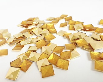 May include: A pile of gold-colored metal pyramid-shaped embellishments. The embellishments are scattered on a white surface.