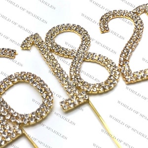 May include: Three gold-colored cake toppers shaped like the numbers 1, 8, and 2. Each number is covered in clear rhinestones and has a gold wire attached to the back for inserting into a cake.