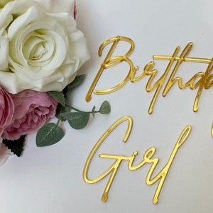 Op de afbeelding: Gouden acryl letters in schrijfletters die "Birthday Girl" spellen op een witte achtergrond met roze en witte bloemen.