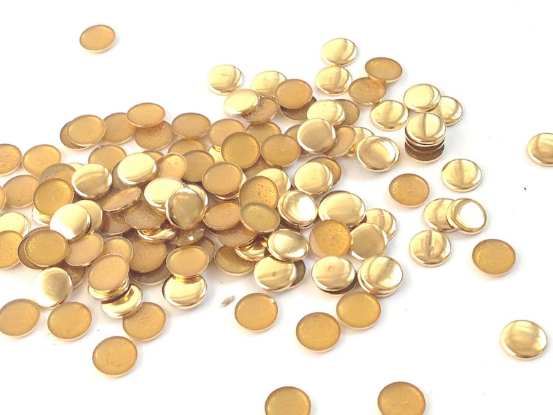 May include: A large pile of gold-colored metal round embellishments. The embellishments are scattered on a white surface.