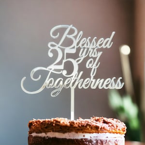 Peut inclure: Un cake topper argenté avec l'inscription "Blessed 25 yrs of Togetherness" en écriture cursive. Le cake topper est placé sur un gâteau à plusieurs couches avec des copeaux de chocolat.