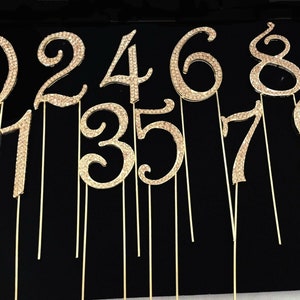 May include: Set of ten gold-coloured cake toppers, each featuring a rhinestone-covered number from 0 to 9. The numbers are attached to thin gold wires.