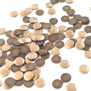 May include: A pile of round, flat, brass-colored metal discs. The discs are scattered on a white surface.