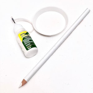 Op de afbeelding: Een wit potlood, een klein flesje Gem-Tac permanente lijm en een wit plastic schaaltje op een wit oppervlak.