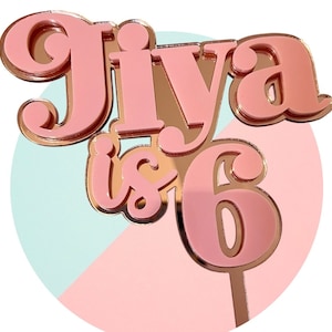Pode incluir: Topo de bolo de acrílico rosa com o nome "Jiya is 6" em uma fonte retrô.