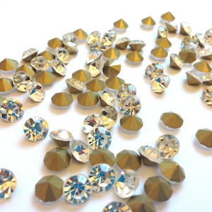 Puede incluir: Una dispersión de diamantes de imitación de vidrio transparente con engastes de metal dorado. Los diamantes de imitación son redondos y tienen un diseño facetado. Los engastes metálicos son planos y tienen una superficie ligeramente texturizada.