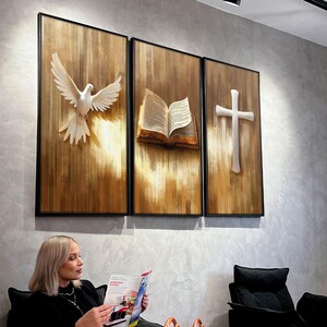 K&ouml;nnte beinhalten: Ein Triptychon aus gerahmten Kunstpaneelen mit religi&ouml;sen Symbolen. Die Paneele zeigen eine wei&szlig;e Taube, ein offenes Buch und ein Kreuz, alles vor einem strukturierten, holzfarbenen Hintergrund. Das Kunstwerk ist in Schwarz gerahmt.