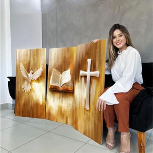 K&ouml;nnte beinhalten: Dreiteilige Wandkunst aus Holz mit religi&ouml;sen Symbolen. Die Paneele zeigen eine wei&szlig;e Taube, ein offenes Buch und ein wei&szlig;es Kreuz. Das Kunstwerk hat ein warmes, goldenes Farbschema mit strukturierten Details.