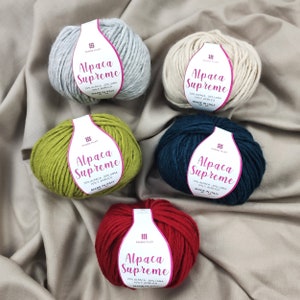 Puede incluir: Cinco ovillos de lana en diferentes colores: gris, beige, verde, azul verdoso y rojo. Cada ovillo tiene una etiqueta que dice "Alpaca Supreme" con el texto "70% Alpaca - 20% Lana - 10% Acrilica" y "Made in Italy".