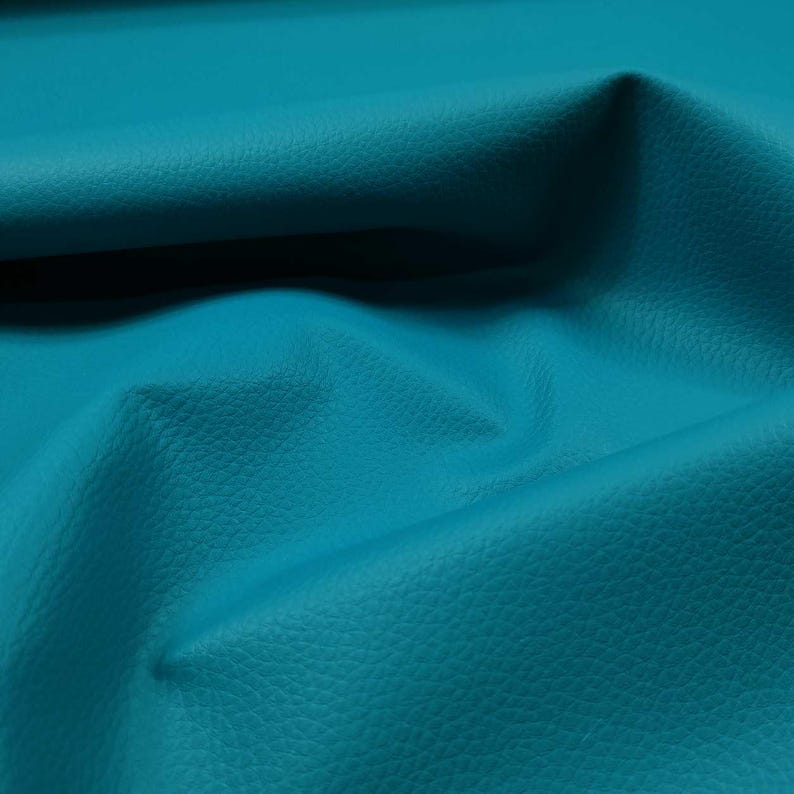 May include: Close-up of a textured, turquoise leather material. The fabric has a subtle grain and is folded, creating shadows and highlights that emphasize its texture and color. The overall appearance is smooth and luxurious.