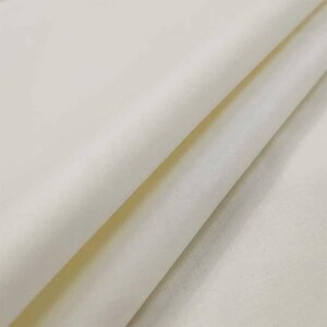 Peut inclure: Gros plan sur un tissu plié blanc cassé. Le tissu a un aspect doux et légèrement texturé avec des plis et des ombres visibles, suggérant un matériau léger comme le lin ou le coton. La couleur est un blanc chaud et crémeux.