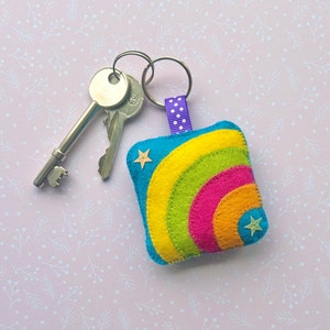 May include: A colourful felt keyring with a rainbow design. The keyring has a blue background with a yellow, green, pink, and orange rainbow. Two small star-shaped embellishments are on the keyring. Two silver keys are next to the keyring.