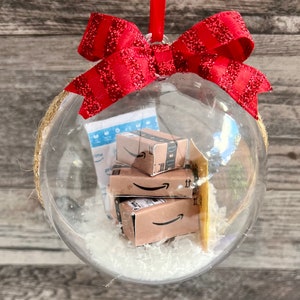 May include: A clear Christmas ornament filled with miniature Amazon boxes and packing material. The ornament is topped with a red glittery bow and a red ribbon for hanging. The ornament is a festive holiday decoration.