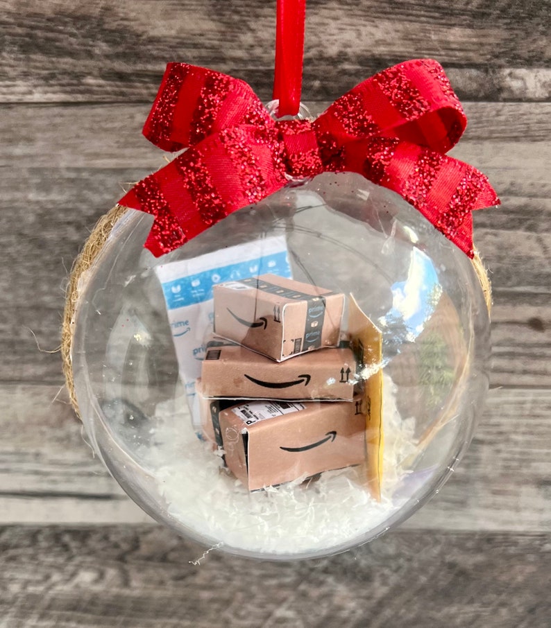 May include: A clear Christmas ornament filled with miniature Amazon boxes and packing material. The ornament is topped with a red glittery bow and a red ribbon for hanging. The ornament is a festive holiday decoration.