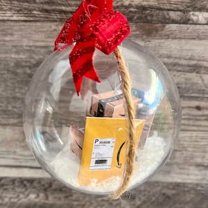 May include: Clear Christmas ornament with miniature Amazon Prime boxes and a shipping label inside. The ornament is decorated with a red glittery bow and a jute twine hanger. The background is a wooden surface.