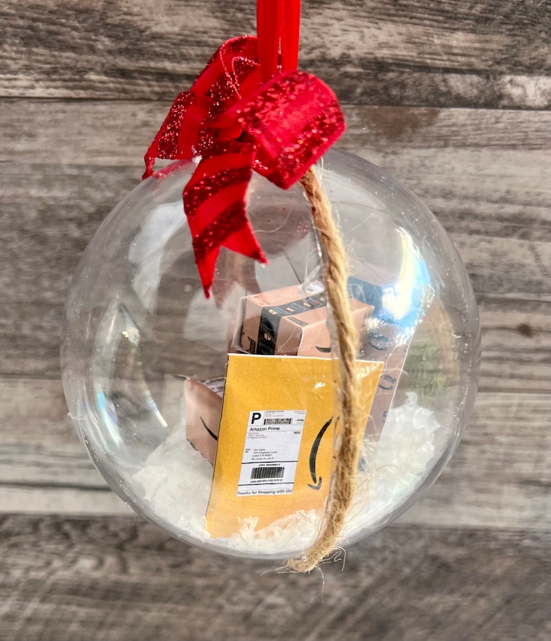 May include: Clear Christmas ornament with miniature Amazon Prime boxes and a shipping label inside. The ornament is decorated with a red glittery bow and a jute twine hanger. The background is a wooden surface.