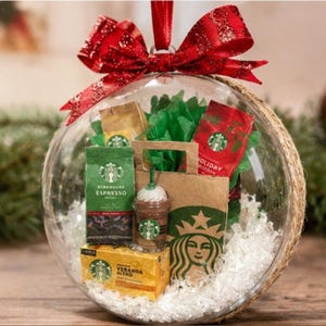 May include: A clear glass ornament with a red bow, filled with Starbucks coffee products. The ornament contains coffee bags, a small paper bag, and a miniature coffee cup. The ornament is decorated with white filler.