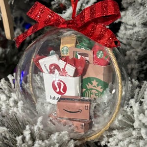 May include: Clear Christmas ornament filled with miniature shopping bags and boxes, including a Starbucks bag, Lululemon bag, and Amazon boxes. The ornament is topped with a red glittery bow and hangs from a gold string.