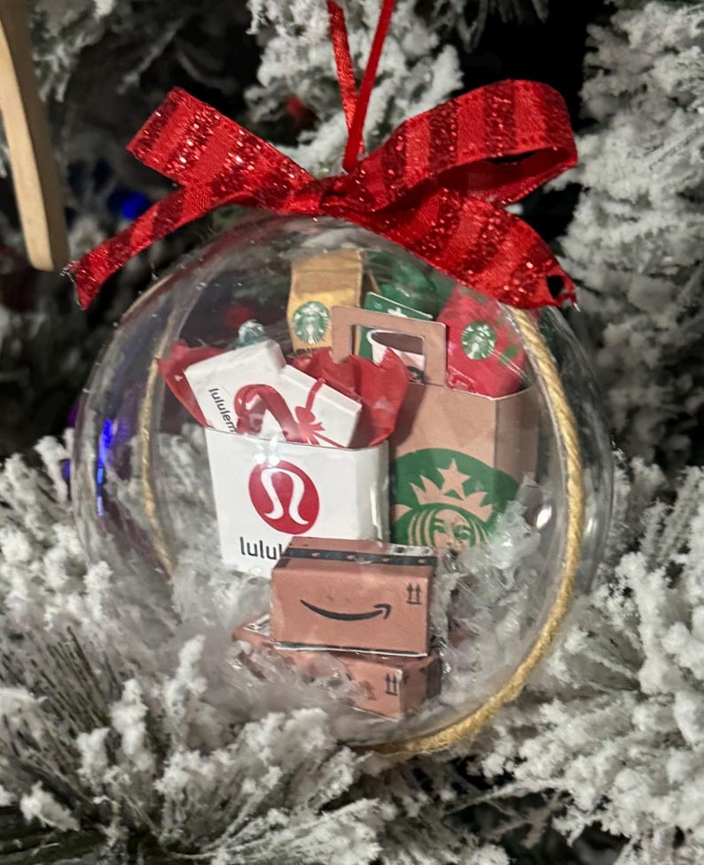 May include: Clear Christmas ornament filled with miniature shopping bags and boxes, including a Starbucks bag, Lululemon bag, and Amazon boxes. The ornament is topped with a red glittery bow and hangs from a gold string.