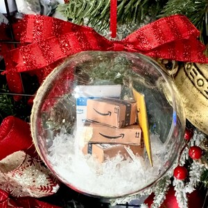 May include: A clear glass Christmas ornament filled with miniature Amazon boxes and white faux snow. The ornament is topped with a large red glittery bow and hangs from a red ribbon. Festive holiday decor.