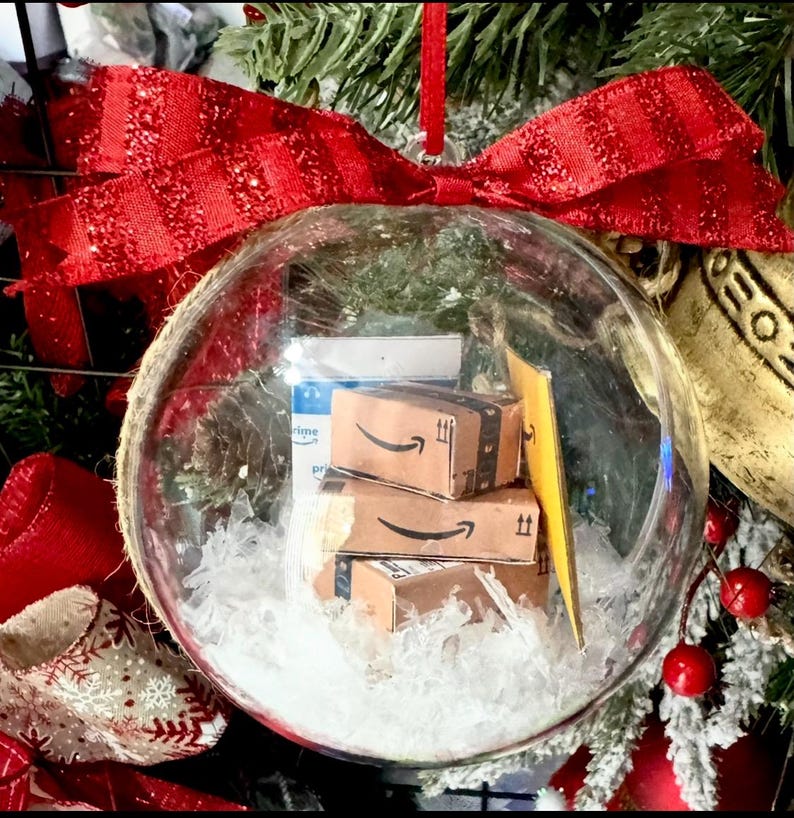 May include: A clear glass Christmas ornament filled with miniature Amazon boxes and white faux snow. The ornament is topped with a large red glittery bow and hangs from a red ribbon. Festive holiday decor.