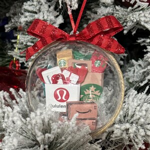 May include: A clear glass Christmas ornament filled with miniature paper shopping bags and gift boxes. The ornament includes a red glittery bow and features logos from Starbucks, Lululemon, and Amazon. The ornament hangs on a snow-covered Christmas tree.