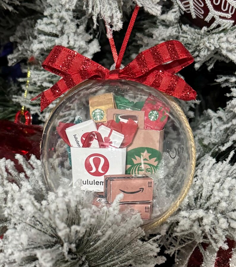May include: A clear glass Christmas ornament filled with miniature paper shopping bags and gift boxes. The ornament includes a red glittery bow and features logos from Starbucks, Lululemon, and Amazon. The ornament hangs on a snow-covered Christmas tree.