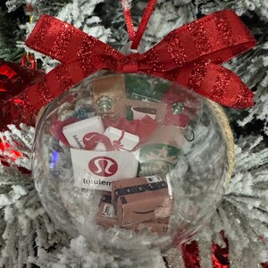 May include: Clear glass Christmas ornament filled with miniature shopping bags and boxes, including a Lululemon bag and an Amazon box. The ornament is topped with a red glittery bow and hangs on a snow-covered Christmas tree.