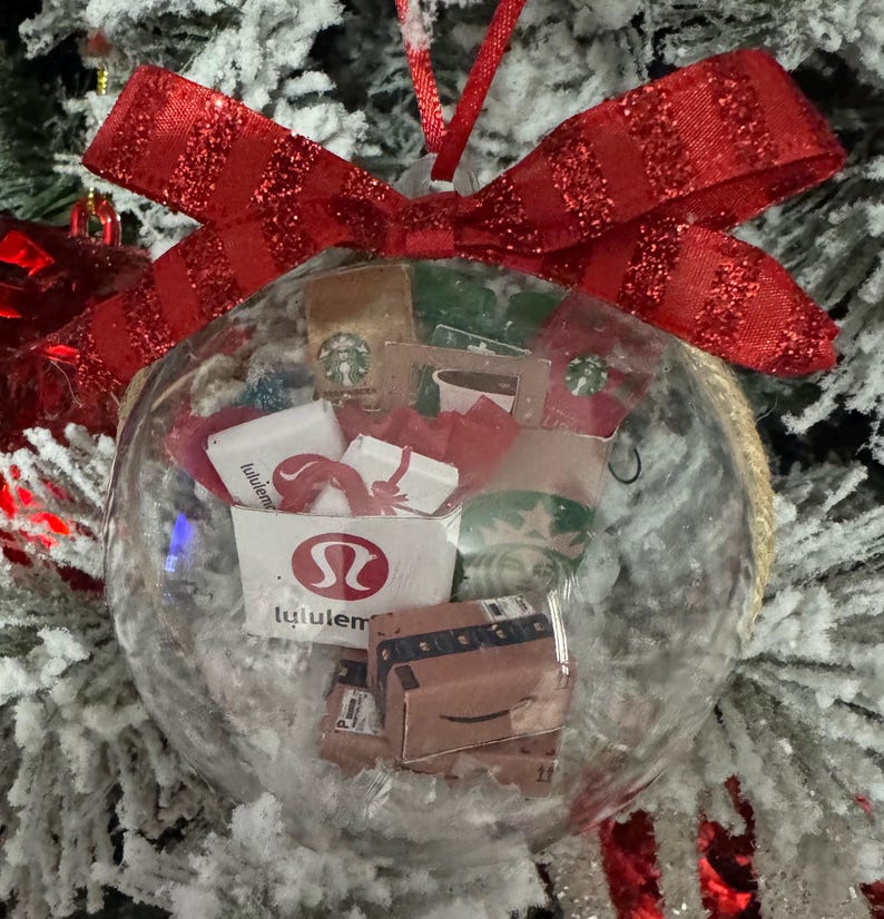 May include: Clear glass Christmas ornament filled with miniature shopping bags and boxes, including a Lululemon bag and an Amazon box. The ornament is topped with a red glittery bow and hangs on a snow-covered Christmas tree.
