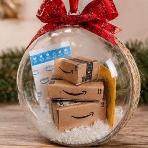 May include: A clear glass Christmas ornament filled with miniature Amazon boxes, a Prime shipping bag, and a yellow envelope. The ornament is topped with a red glittery bow and hangs from a red ribbon. The ornament is sitting on a wooden surface.