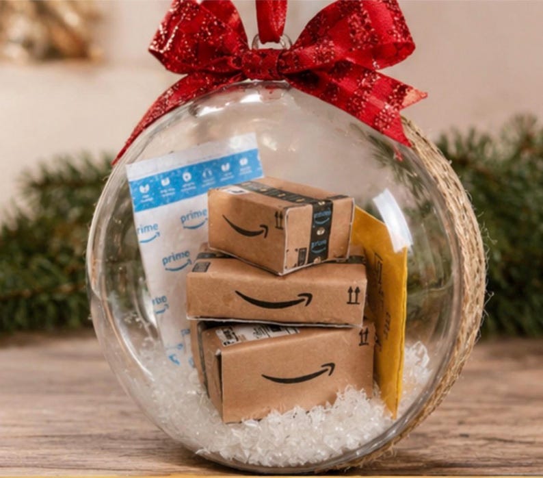 May include: A clear glass Christmas ornament filled with miniature Amazon boxes, a Prime shipping bag, and a yellow envelope. The ornament is topped with a red glittery bow and hangs from a red ribbon. The ornament is sitting on a wooden surface.